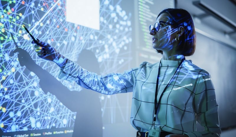 Image for Young Female Teacher Giving a Data Science Presentation in a Dark Auditorium with Projecting Slideshow with Artificial Intelligence Neural Network Architecture. Taking a people first approach to AI implementationConcept stock photo