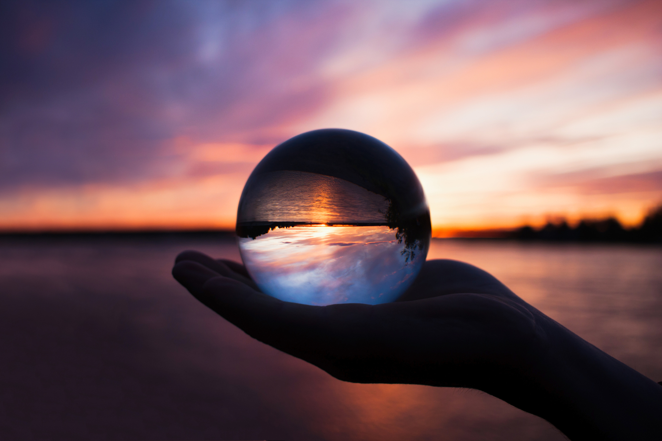 Beautiful scenic view through lens of crystal ball on water horizon at summer sunset. Clouds reflection in sphere. Palm of hand holding globe in outdoor. stock photo